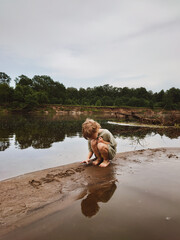 Child playing in wet sand by a quiet river on a cloudy day, surrounded by green forest. Peaceful nature scene with reflection in water. Perfect for themes of childhood, freedom, and nature.
