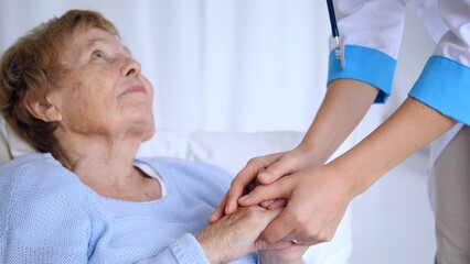 Young doctor holding the hands of an elderly patient lying in bed, providing comfort and support in...