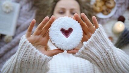 Cozy picnic scene with woman wearing a white knitted sweater, holding a heart shaped biscuit covered in powdered sugar, with blurred background featuring book, cookies, candle and lavender