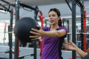 Asian sportswoman doing fitness ball exercise with personal trainer in gym