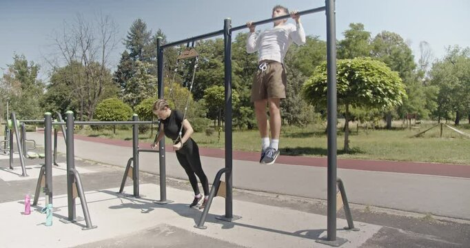 A young, fit couple exercising together on a sunny day at an outdoor gym. They are doing pull-ups and triceps extensions, enjoying the benefits of fresh air and sunshine.