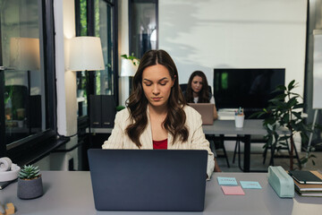 Young businesswoman working on laptop in modern office with colleagues in background