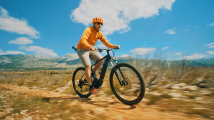 A man, wearing a yellow helmet and shirt, rides an e-bike on a dirt trail. The camera captures a dynamic, fast-paced shot.