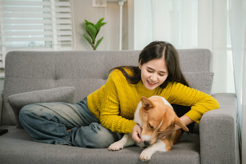 A young woman relaxes indoors on a cozy sofa, smiling as she bonds with her playful dog, enjoying...