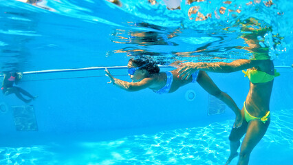 A woman in a yellow bikini teaches a young girl in a pool, demonstrating swimming techniques...