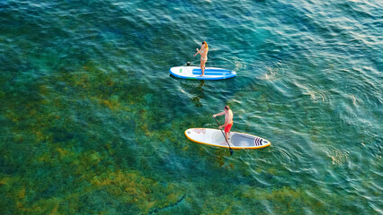 Aerial view of two people paddleboarding on clear blue water, enjoying an active aquatic sport.