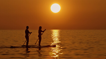 A stunning sunset paddleboard scene featuring two people enjoying the tranquil waters. The warm hues of the setting sun create a serene and picturesque atmosphere. Aerial view.