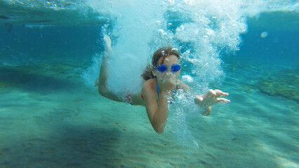 Woman of Asian descent swims underwater in a pool, wearing blue goggles. Underwater view captures her swimming motion and the bubbles around her.