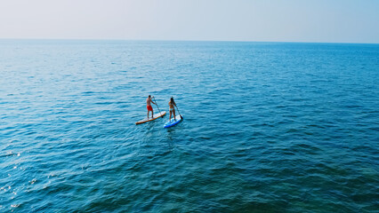An aerial view captures two people paddleboarding on a vast, blue ocean. They stand on surfboards,...