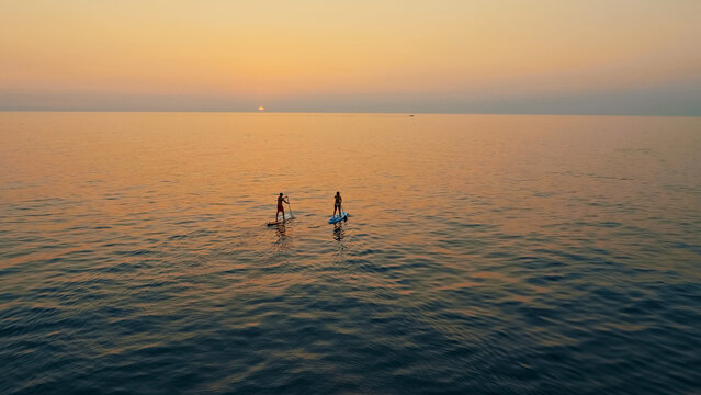 Aerial view captures two people paddleboarding on the calm ocean at sunset. Warm hues of the sky reflect on the water, creating a serene and tranquil atmosphere.