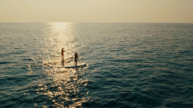 Breathtaking aerial view showcases two people paddleboarding on the ocean, their silhouettes beautifully contrasting against the shimmering water. Scene radiates tranquility and freedom.