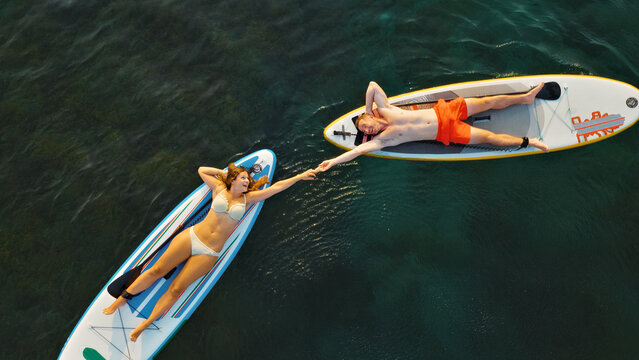 An aerial view captures a man and a woman paddleboarding on a serene lake, holding hands while lying on their boards. The shot emphasizes their connection and enjoyment of the water sport. - Powered by Adobe