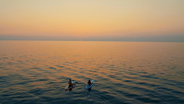 A serene sunset captures two people paddleboarding on the calm ocean, enjoying the tranquil waters and the warm hues of the setting sun. Aerial view.