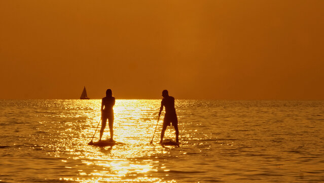 A silhouette of a man and a woman paddleboarding on a golden sunset, exuding tranquility and adventure. Aerial view.