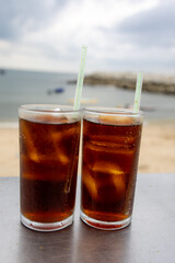 glass of iced cola sits on a table by the sea, with a cloudy sky backdrop. The drink's bubbles and condensation are visible