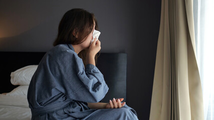 Woman holds throat in discomfort sitting on edge of bed and sneezes
