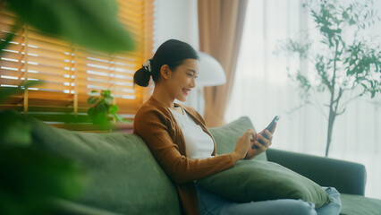 Smiling pretty young Asian woman sitting on cozy couch, using funny mobile apps in living room. Woman at home, Doing online shopping, Messaging friends, Posting on social Media