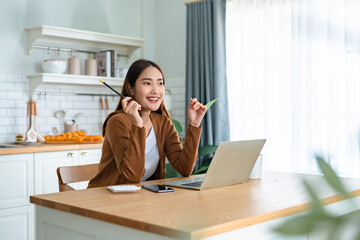 Beautiful young Asian woman working with laptop computer in home kitchen. Work at home