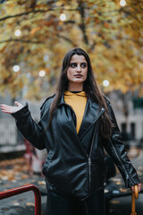 A woman dressed in a black leather jacket and yellow top poses confidently under vibrant fall foliage adorned with string lights, providing a casual and stylish urban setting.
