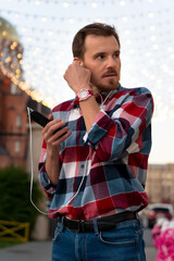 Man adjusting earphones while listening to music under lights. Guy with beard and modern hairstyle wearing in blue, red and white plaid shirt walking through the city streets in urban background