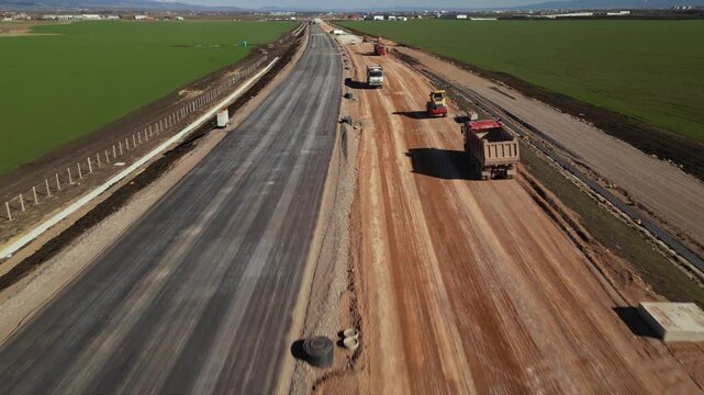 Aerial view of dump trucks and construction vehicles moving along a dirt haul road at a large earthworks site, with cut embankments, open fields, and sunset light over the landscape