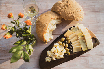 Tasty cheese and olives on a wooden board and homemade bread on the table on a sunny day in golden hour and a glass of wine and flower decoration