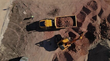 Top-down aerial drone view of a yellow dump truck being loaded with soil by an excavator on a dusty construction site, with rough ground textures, tire tracks, and a worker in a safety vest highlighti