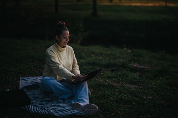 Obraz premium A girl sits on a blanket in the evening, working on a laptop outdoors.