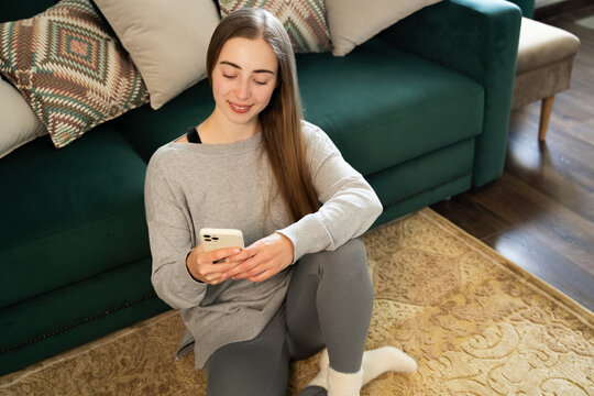 Young smiling woman using smartphone holding cellphone looking at mobile screen texting relaxing wear casual clothes sitting on floor in living room at home, digital communication indoor concept