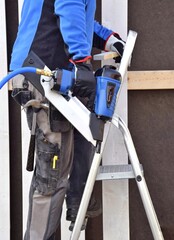 A man stands on a stepladder and holds a pneumatic stapler, a nail gun for a wooden board, hammers metal nails and staples in his hand.