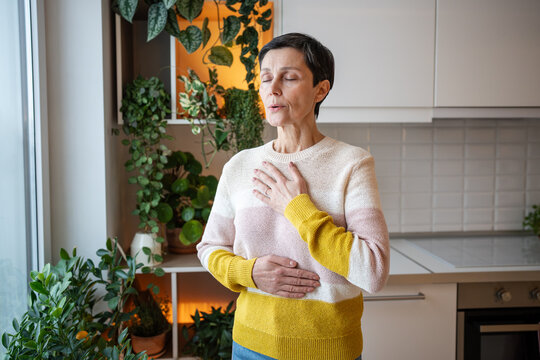 Content serene older woman practicing deep breaths in meditation, hands on torso at home. Stress relief through controlled breathing, comfortable leisure activity, holistic wellness, mindful happiness