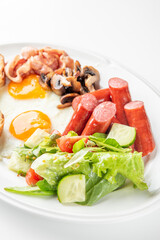 A classic breakfast plate with sunny-side-up eggs, crispy bacon, grilled sausages, sautéed mushrooms, fresh salad with cucumbers and cherry tomatoes, and toasted bread slices, presented on white dish.