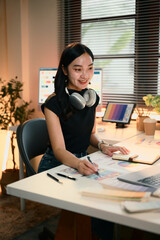 Graphic designer woman is sitting at a desk with a laptop and a notebook. She is wearing headphones and smiling