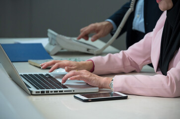Close up shot of woman in a pink blazer and black hijab typing on  laptop with smartphone on office desk while businessman using landline phone in background