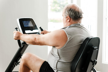 Overweight senior man in sleeveless shirt exercising on a stationary recumbent bike indoors, focusing on fitness, health improvement, and active aging in a bright wellness-focused environment.