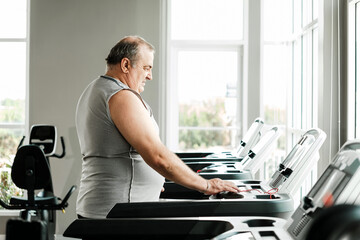 Overweight senior man preparing to use a treadmill in a bright gym, promoting wellness and a healthy lifestyle. Focused on fitness, aging healthily, and improving personal well-being.