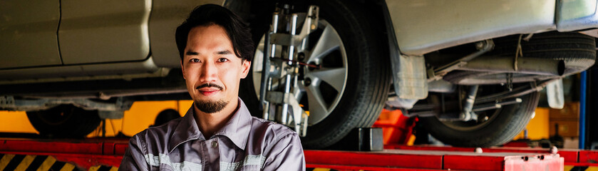Confident auto mechanic in gray uniform stands with arms crossed in front of elevated vehicle at repair shop, representing expert service, professional inspection, and car maintenance confidence.