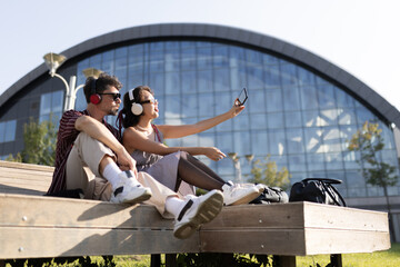Young couple sitting on a bench with headphones, taking a selfie outside modern architectural building on a sunny day