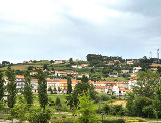Scenic view of a hillside town, showcasing residential architecture nestled among green trees and lush landscapes under a cloudy sky. Ideal for travel, real estate, or lifestyle themes.