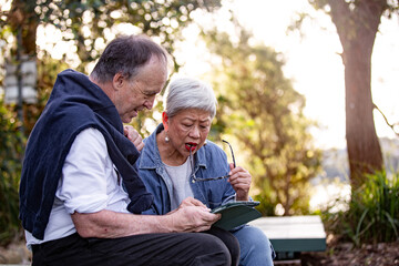 Senior couple sitting at park bench looking at computer tablet