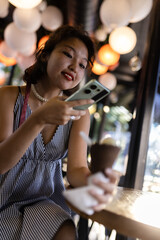 Smiling young woman enjoying ice cream while using smartphone in a lively cafe with glowing lights