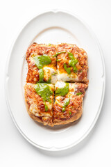 Close-up of a crispy Italian-style pizza bread topped with melted cheese, tomato sauce, and green pesto, served on a white plate. The vibrant colors and textures make it look appetizing and fresh
