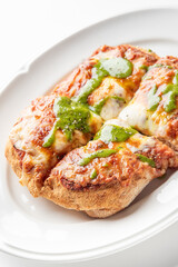Close-up of a crispy Italian-style pizza bread topped with melted cheese, tomato sauce, and green pesto, served on a white plate. The vibrant colors and textures make it look appetizing and fresh
