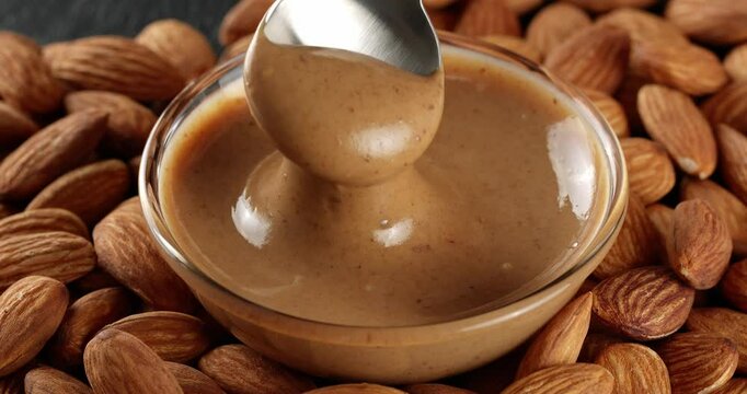Almond nut butter is being scooped with a spoon. The almond butter drips from the spoon into a glass bowl on a background of almond nuts