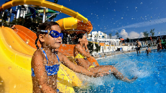 A close-up shot of two young girls splashing down a yellow water slide into a pool, wearing goggles and swimsuits, enjoying a fun summer day at an aqua park.
