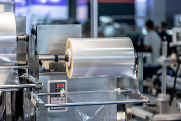 Industrial machine with roll of transparent plastic film on metal stand in factory. Automated equipment for packing goods. Focus on equipment. Selected focus