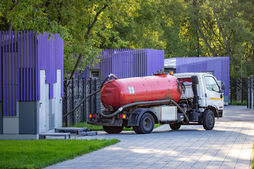 Vacuum tanker truck emptying septic tank on residential street, red tank and white cab, trees and modern fencing, sunny day, urban maintenance, environmental service