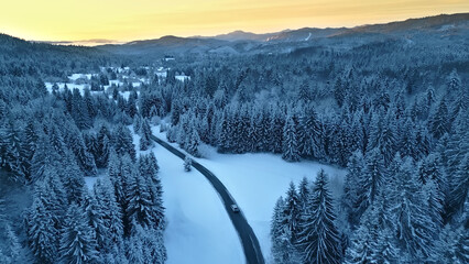 An aerial view of a winding road through a snowy forest, with a car driving along the route. The...