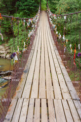 Wooden suspension bridge with colorful ribbons hanging from wires crosses fast mountain river, mountain creek in lush green coniferous Carpathian forest. Adventure, travel, hiking and local charm.