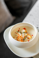 A gourmet soup served in a white bowl, garnished with shredded cheese, croutons, fresh herbs, and drops of red sauce. The creamy texture and artistic presentation make it a visually appealing dish